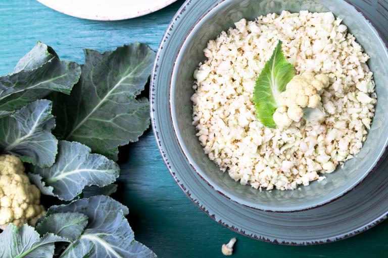 Raw Cauliflower Rice Cauliflower rice in a blue bowl on a blue table next to a head of cauliflower with leaves.